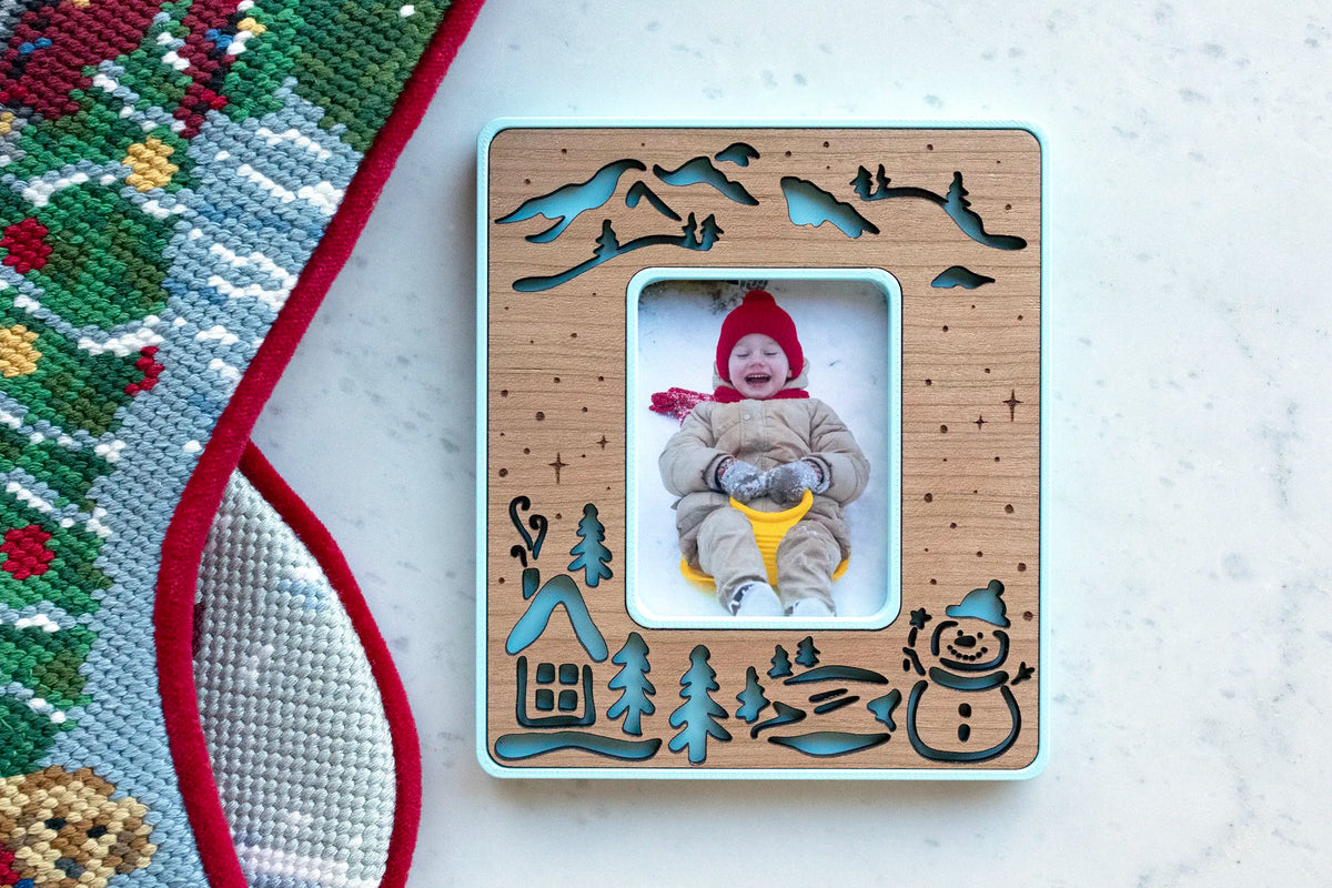 Ice blue snowman magnetic photo frame with a cherry wood inlay of a snowy cabin, pine trees, mountains, and a snowman, holding a photo of a toddler in a red hat sledding, beside a Christmas stocking.