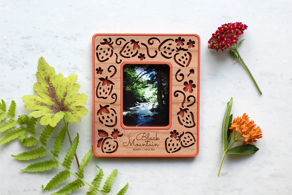 A magnetic strawberry polaroid photo frame made with a plastic housing and laser cut wooden inlay. The strawberry frame has a whimsical, laser cut design and a red border. The words "Black Mountain North Carolina" are engraved in the frame for personalization. Flowers are lying around the frame.