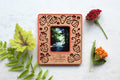 A magnetic strawberry polaroid photo frame made with a plastic housing and laser cut wooden inlay. The strawberry frame has a whimsical, laser cut design and a red border. The words "Black Mountain North Carolina" are engraved in the frame for personalization. Flowers are lying around the frame.