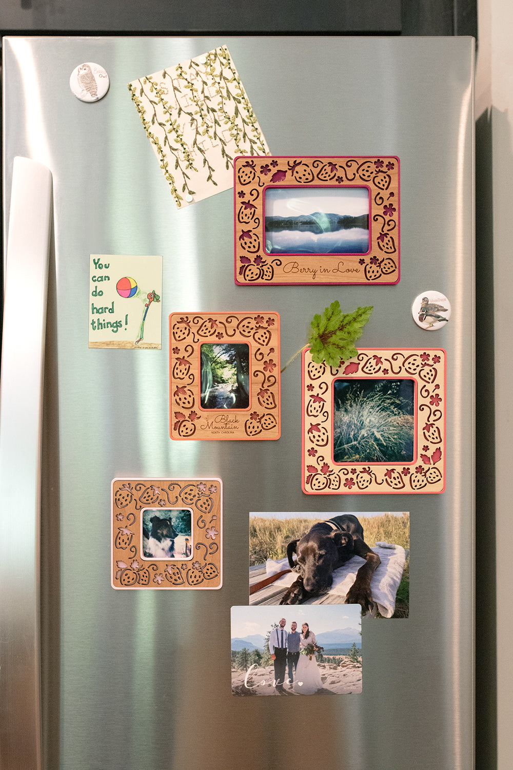 An assortment of four whimsical, laser cut strawberry Polaroid photo frames in various sizes, colors, and wood types. The magnetic frames are mounted to the front of a refrigerator and displaying a Polaroid photo, a Fujifilm Instax Mini photo, an Instax Wide photo, and a Polaroid Go photo.