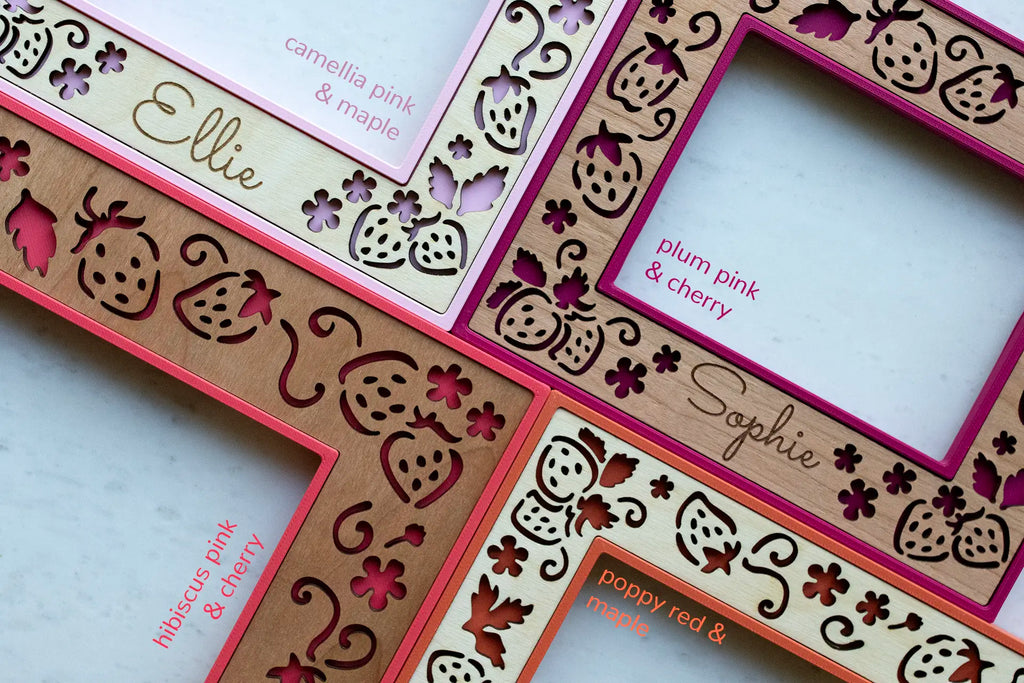 Close-up of four colorful, laser cut, strawberry frames with various wooden inlays showing the grain and finish of the wood. Frames are camellia pink, plum pink, hibiscus pink, and poppy red. Wood inlay options are cherry, walnut, and maple.