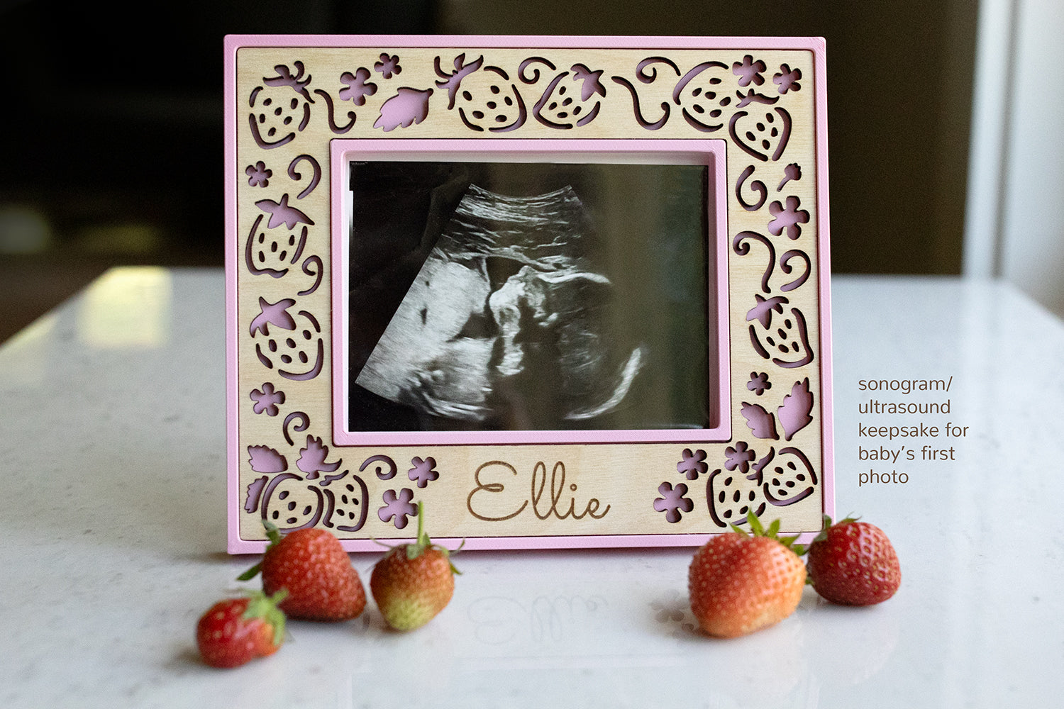 An ultrasound frame in 3" x 4" to fit standard ultrasound / sonogram photographs. Frame is light pink with a laser cut birch wood inlay and personalization. The birch inlay features strawberries and the name "Ellie" at the bottom. Real strawberries are in the foreground.
