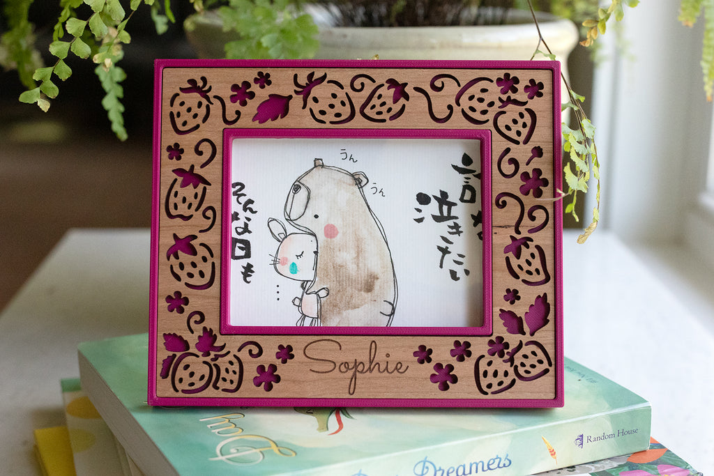 An ultrasound frame featuring the personalized name "Sophie" laser engraved in cherry wood. The frame features laser cut strawberries and is a dark pink color called "plum pink". The small frame sits on a pile of children's books.