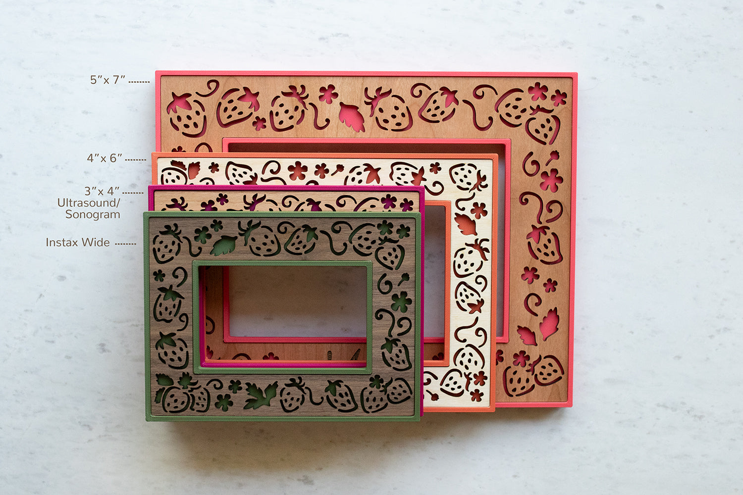 Top view of four frame sizes stacked in a pile on a white marble surface. Strawberry frames come in four sizes: 5"x7", 4"x6", 3"x4" standard ultrasound / sonogram size, and Fujifilm Instax Wide Polaroid film. The frames are whimsical and playful in design.