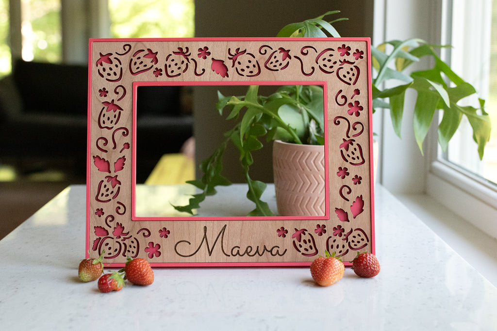 A whimsical strawberry frame featuring a bright pink frame and detailed laser cut strawberries in a custom cherry wooden inlay. Frames are available in 5x7, 4x6, Polaroid Instax Wide, and ultrasound frame sizes. The frame reads "Maeva" at the bottom and has strawberries next to it.