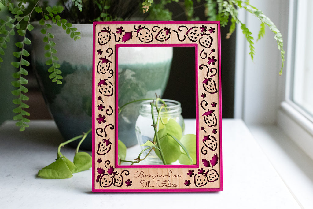 Strawberry picture frame with personalized engraving at the bottom. The wood engraving reads "Berry in Love, The Felixs". The frame housing is a vibrant fuscia color called "plum pink". There is greenery in the background and natural sunlight in the window.