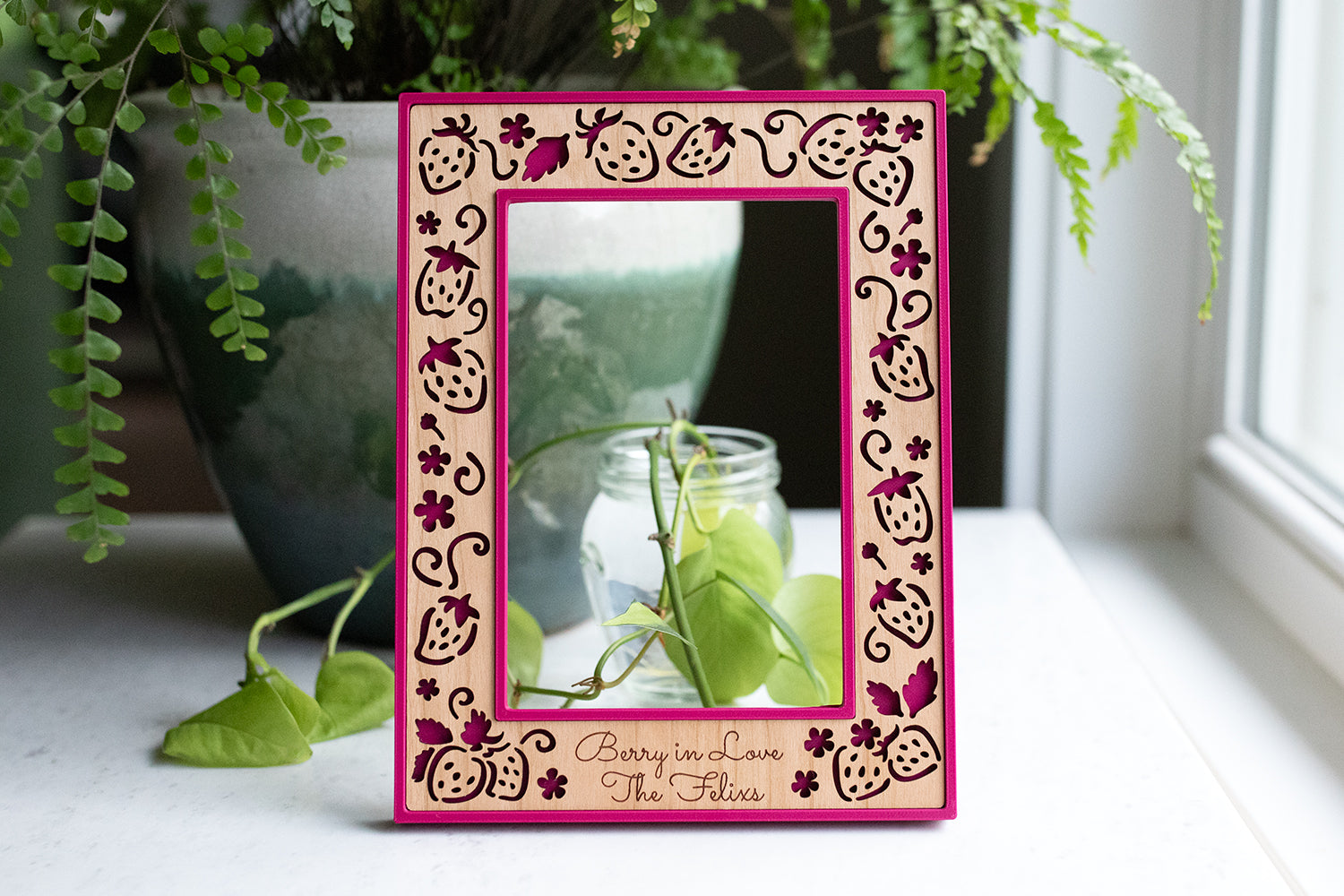 Strawberry picture frame with personalized engraving at the bottom. The wood engraving reads "Berry in Love, The Felixs". The frame housing is a vibrant fuscia color called "plum pink". There is greenery in the background and natural sunlight in the window.