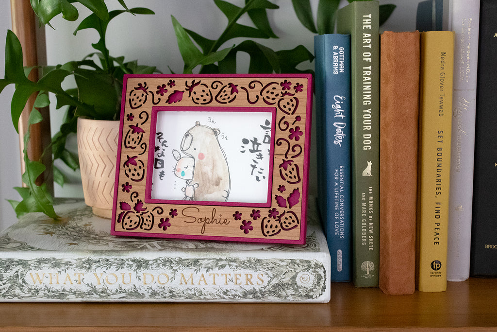 An ultrasound frame with laser cut strawberries and the name "Sophie" laser engraved on a cherry wood inlay. The frame is sitting on a wooden bookshelf. There is greenery in the background and several colorful books next to the frame. The frame displays an illustration of a bear and a bunny hugging with Japanese characters.