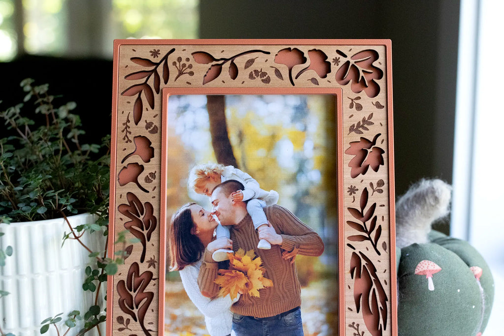 Close-up of a terracotta fall picture frame with a laser-cut cherry wood inlay — autumn leaves, acorns, and branches — framing a fall family portrait of a couple with their child in the autumn woods. Styled with a fabric pumpkin and trailing greenery: a handmade autumn keepsake for displaying your seasonal fall family photos.