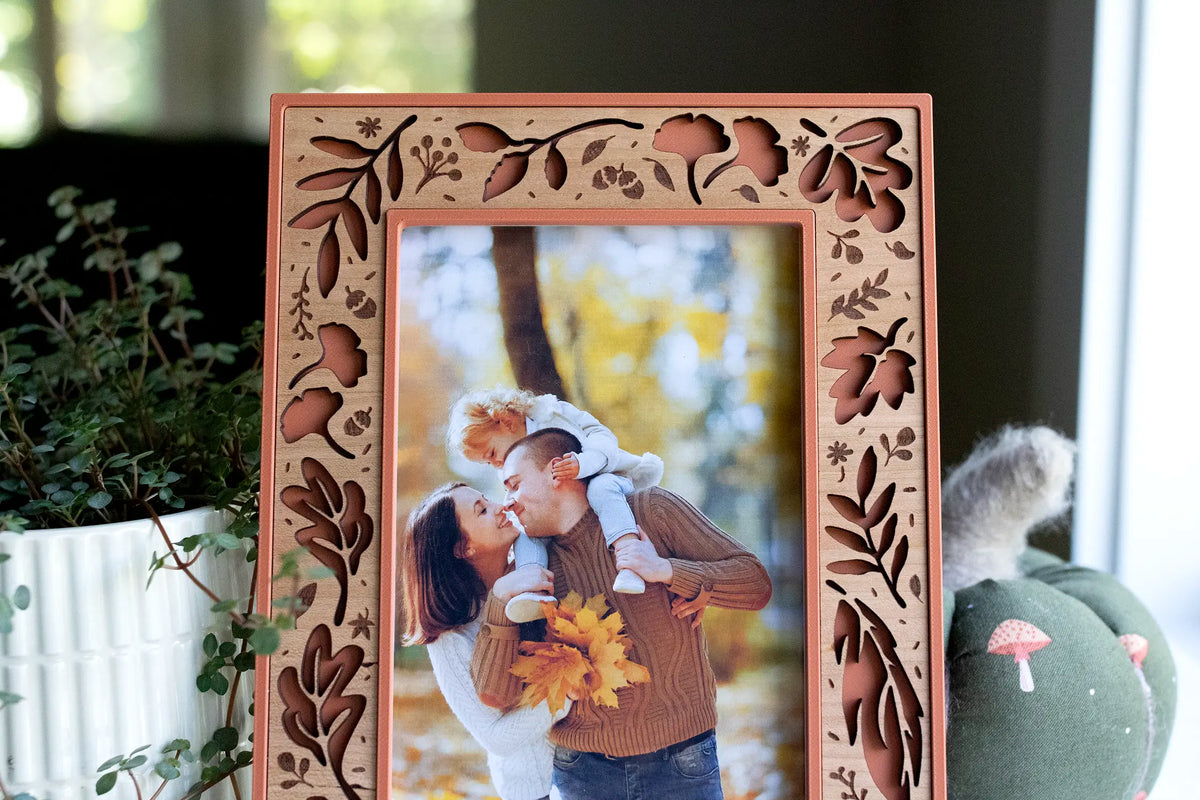 Close-up of a terracotta fall picture frame with a laser-cut cherry wood inlay — autumn leaves, acorns, and branches — framing a fall family portrait of a couple with their child in the autumn woods. Styled with a fabric pumpkin and trailing greenery: a handmade autumn keepsake for displaying your seasonal fall family photos.