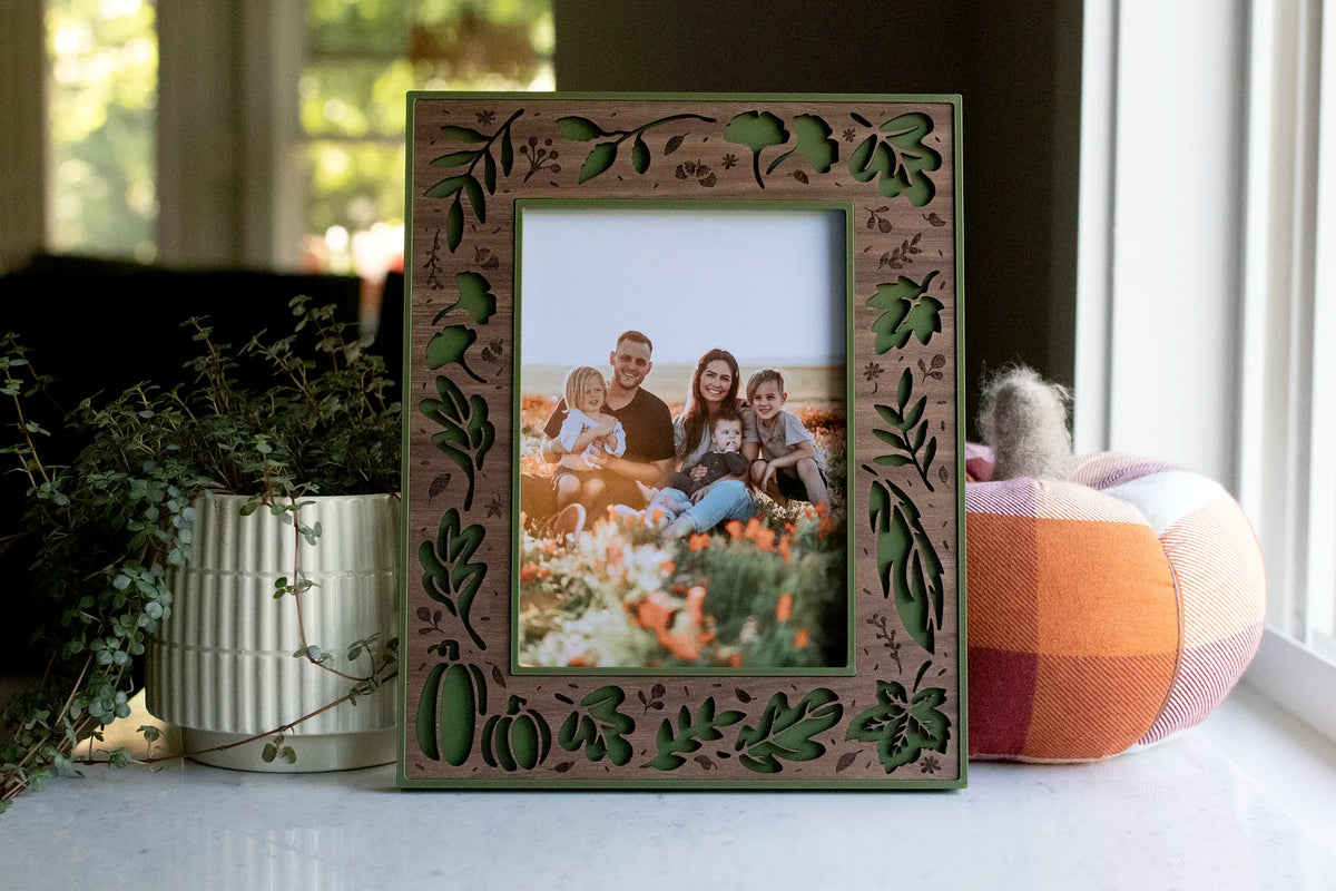 Moss green fall picture frame with a laser-cut walnut wood inlay — oak and maple leaves, pumpkins, acorns, and branches — holding a seasonal fall family portrait taken in a fall flower field. Displayed on a sunny windowsill with a plaid pumpkin and trailing greenery: a handmade autumn keepsake and Thanksgiving decor piece.
