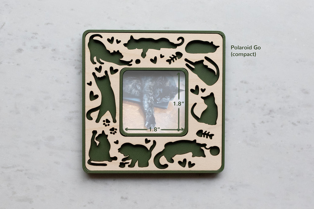 A moss green magnetic cat photo frame in compact Polaroid Go size. The frame features playful laser cut cats amongst little hearts, fish bones, and kitty paw prints. The frame has a wooden inlay cut from maple. The photo opening in the frame is 1.8" x 1.8".