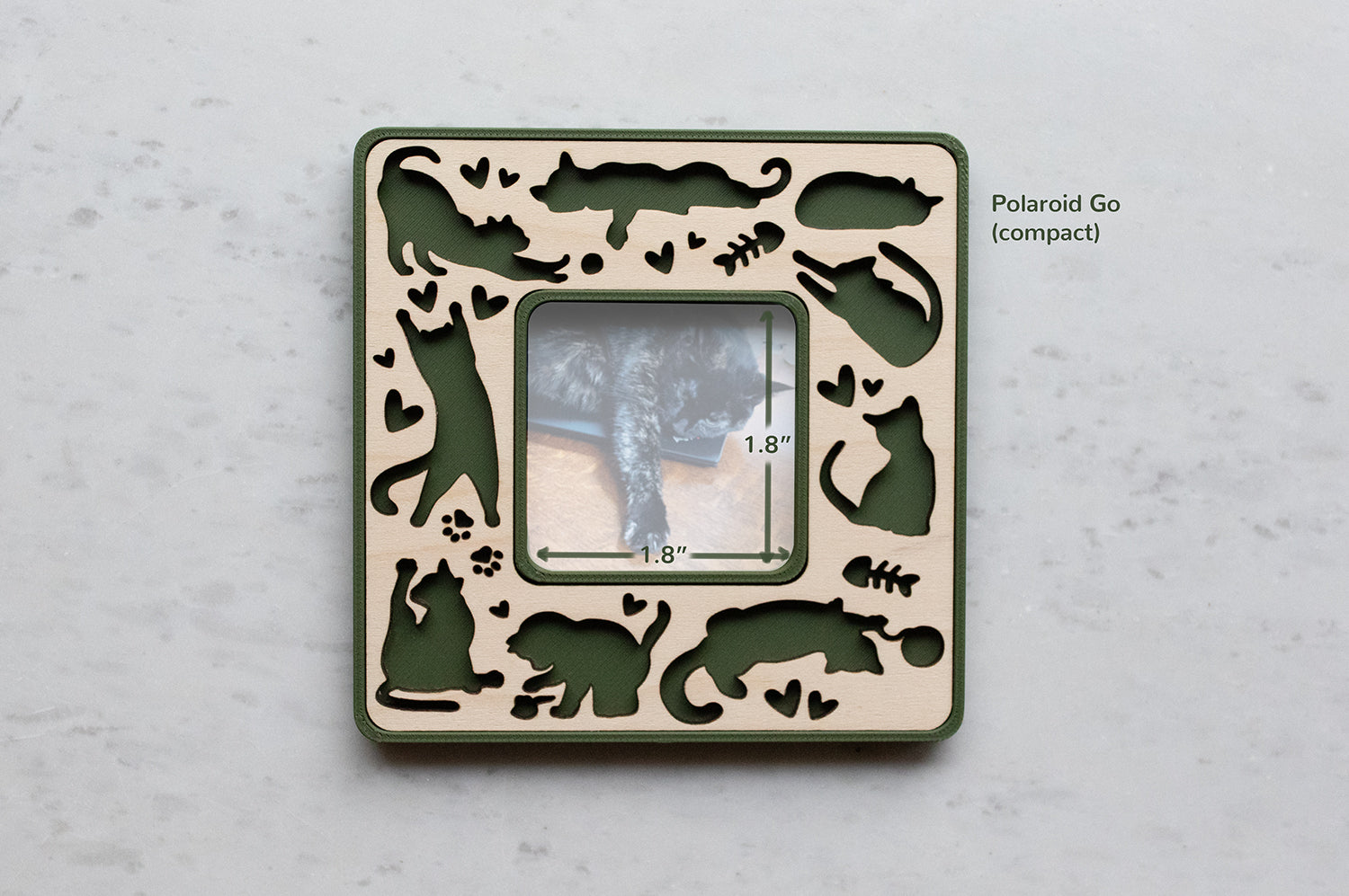 A moss green magnetic cat photo frame in compact Polaroid Go size. The frame features playful laser cut cats amongst little hearts, fish bones, and kitty paw prints. The frame has a wooden inlay cut from maple. The photo opening in the frame is 1.8" x 1.8".