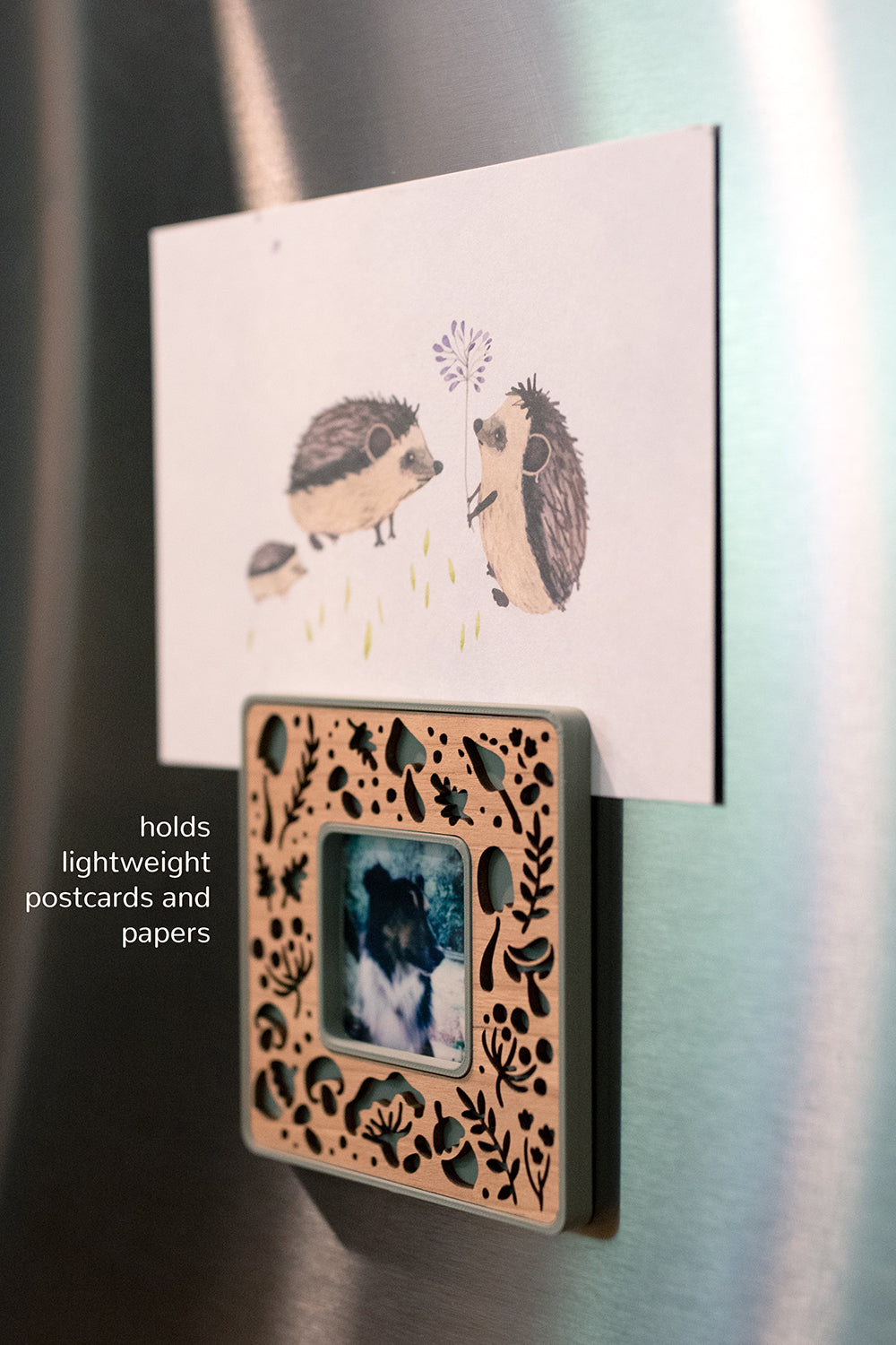 A close-up detail view of a mini Polaroid Go frame with laser cut mushrooms and hanging on a stainless steel refrigerator. The frame is holding up a postcard featuring a cute hedgehog family. The image reads "holds lightweight postcards and papers." The frame is magnetic and includes two neodymium magnets. It is a lightweight frame and can hang on any magnetic surface.