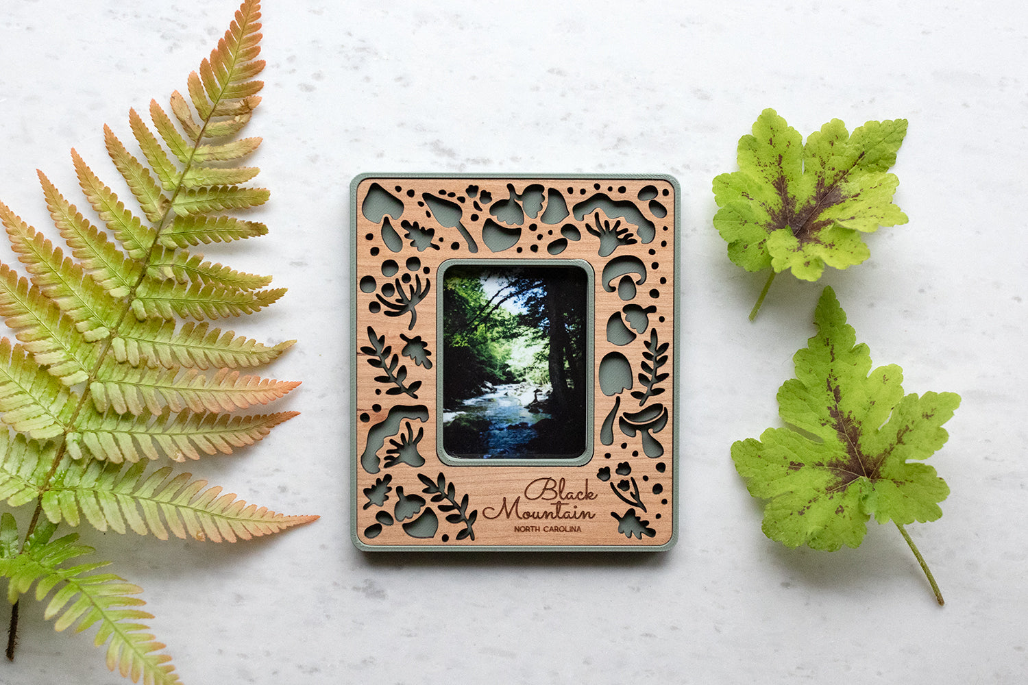 A natural green whimsical mushroom magnetic frame lying next to a large green fern frond. The frame features a delicate pattern of various mushroom types and acorns, including amanita mushrooms, porcini mushrooms, and the classic toadstool mushroom design.