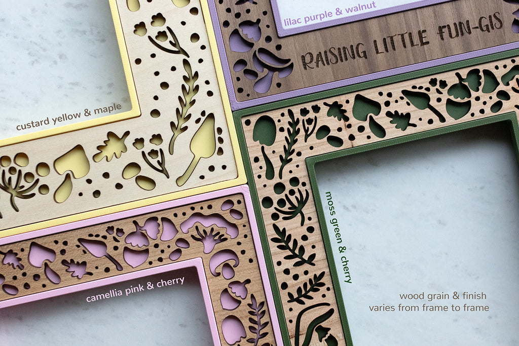 Wooden frames with laser-cut mushroom and acorn designs in various color combinations, including lilac purple with walnut, camellia pink with cherry, moss green with cherry, and custard yellow with maple. There is note that reads "wood grain and finish varies from frame to frame."