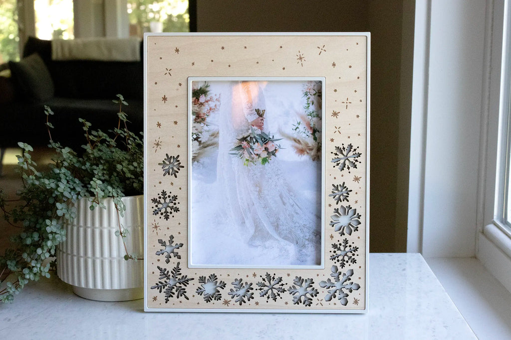 A winter wedding keepsake frame featuring delicate laser-cut falling snowflakes in a maple wood inlay. The frame housing is linen white and displays a photograph of a bride holding her bouquet in the snow at her wedding.