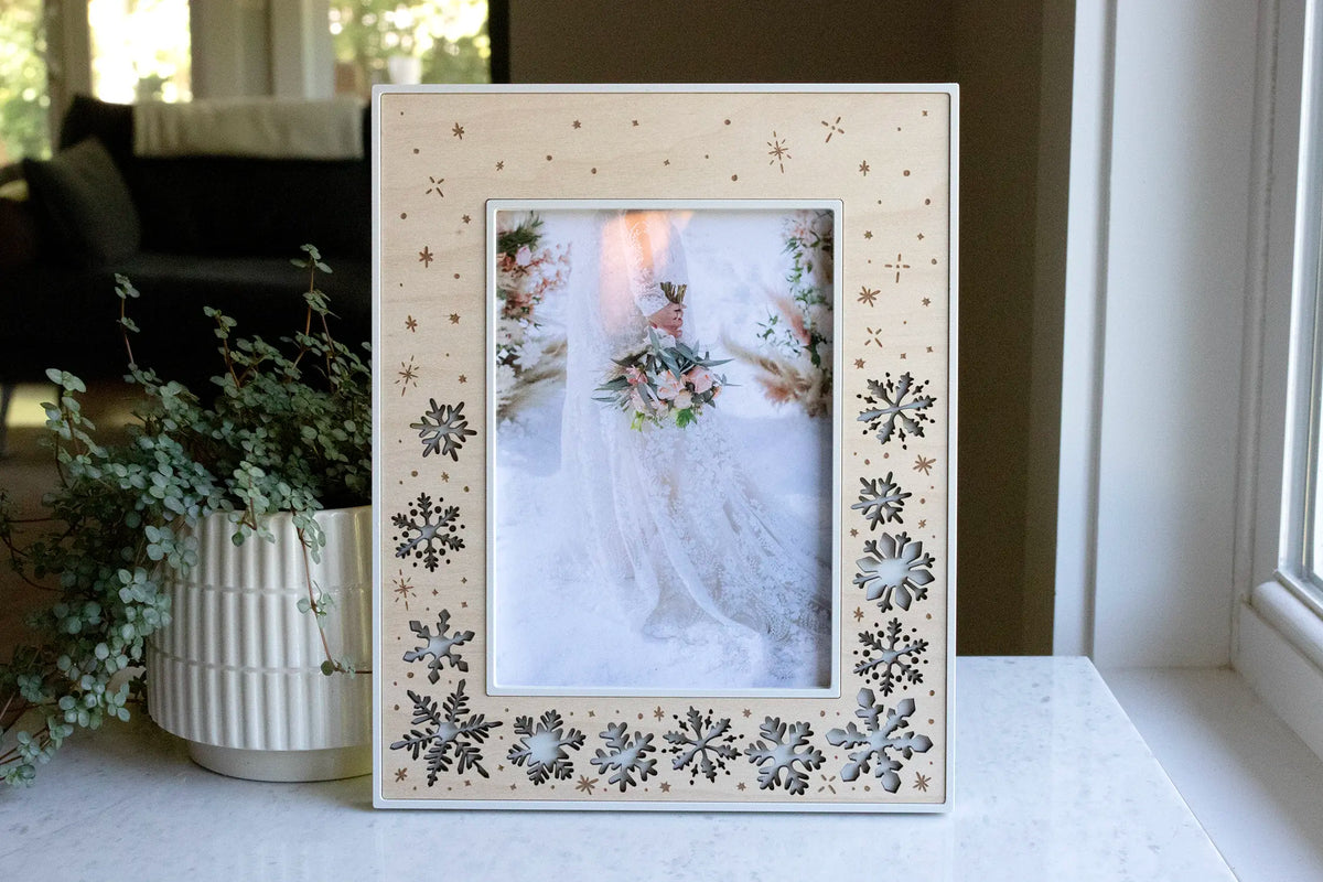 A winter wedding keepsake frame featuring delicate laser-cut falling snowflakes in a maple wood inlay. The frame housing is linen white and displays a photograph of a bride holding her bouquet in the snow at her wedding.