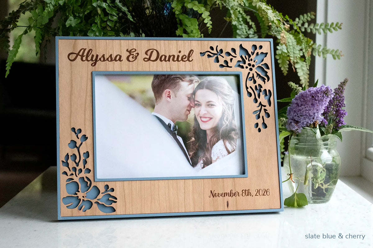 Personalized wedding keepsake frame displaying a photo of newlyweds at their wedding. The frame is slate blue with a cherry wood inlay and engraved with the couple's names "Alyssa & Daniel" and wedding date, "November 8th, 2026".