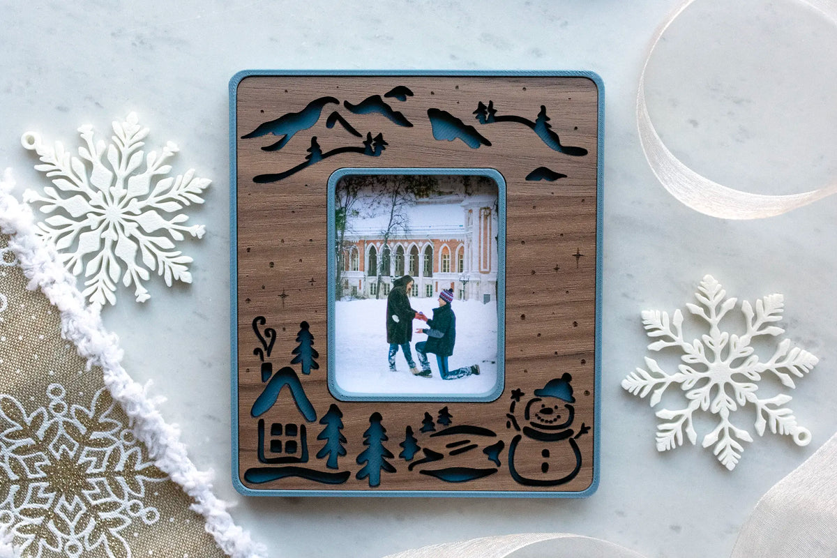 Slate blue and walnut snowman magnetic frame holding a winter engagement photo of a man kneeling to propose in the snow, styled with white snowflake ornaments.