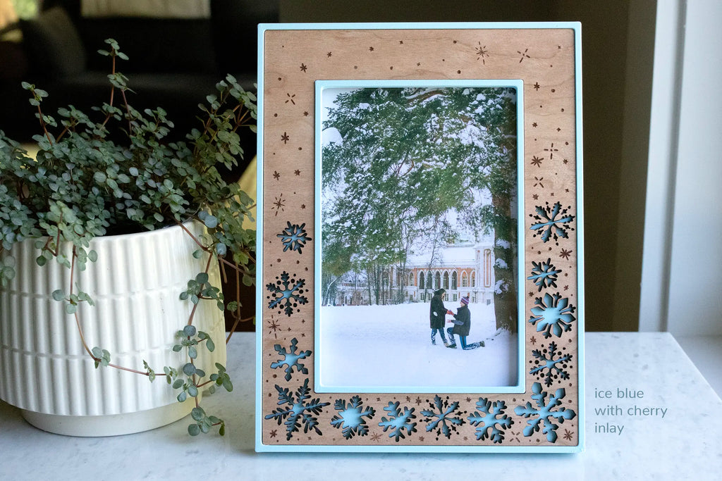 A laser-cut snowflake picture frame with a cherry wood inlay and ice blue housing, holding a photo of a winter proposal — a man kneeling in the snow in a snowy park. Styled on a marble counter beside a eucalyptus plant, it makes a winter engagement keepsake and gift.