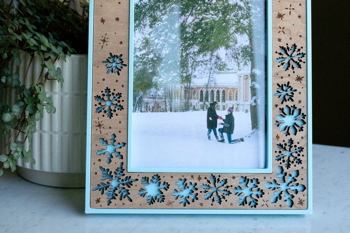 A close-up of a laser-cut snowflake picture frame in ice blue and cherry wood inlay, showing the layered blue snowflake detail around a winter proposal photo — a couple in the snow, one down on one knee. A handmade winter engagement keepsake.