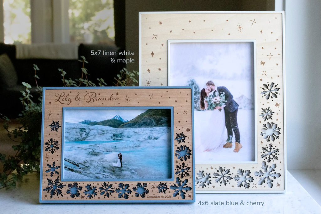 Winter wedding photo frames in two sizes, including 4x6 and 5x7. The 4x6 wedding frame is engraved with the newlyweds names "Lily & Brandon", and their wedding date "December 19, 2026" is engraved in small letters in the bottom right corner of the frame. The larger 5x7 frame features a linen white housing with laser-cut maple wood inlay, and it displays a couple kissing at their winter wedding.