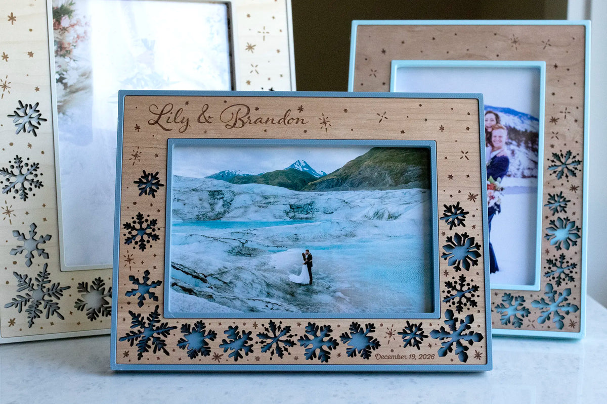 A personalized winter wedding picture frame engraved with the couple's names "Lily & Brandon" and their wedding date, holding a glacier wedding photo, with two more snowflake frames behind it. A handmade laser-cut wedding keepsake and gift idea for newlyweds, in a cherry wood inlay with slate blue housing.
