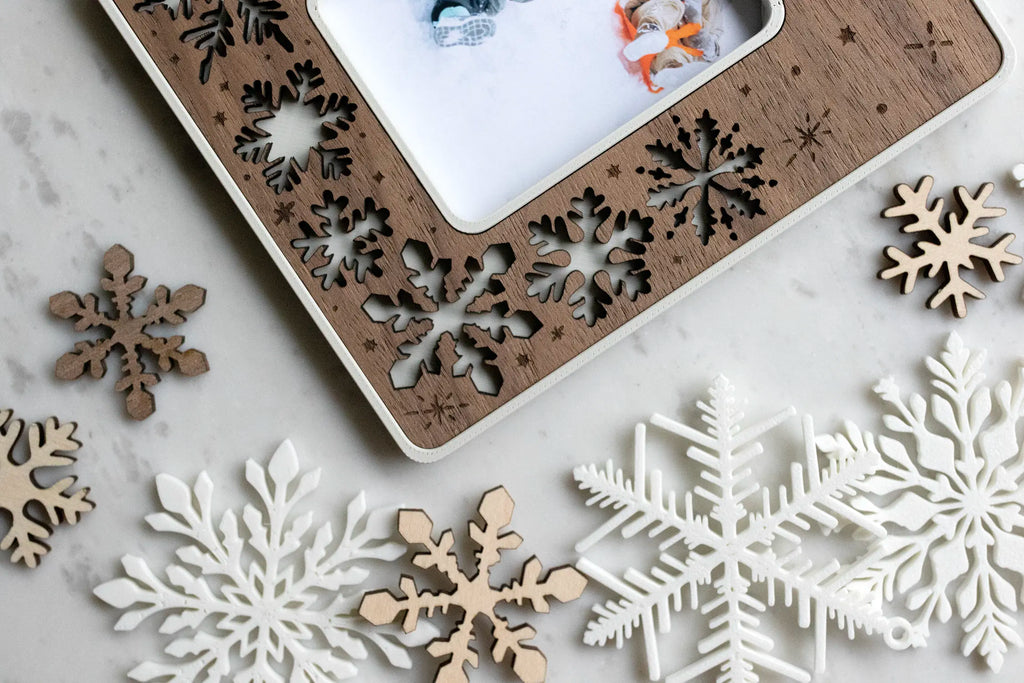 Close-up corner of a laser-cut walnut snowflake magnetic picture frame on white marble, surrounded by scattered wood and white snowflake cutouts, framing a small photo. This holiday keepsake frame is magnetic to stick on the fridge or any magnetic surface.