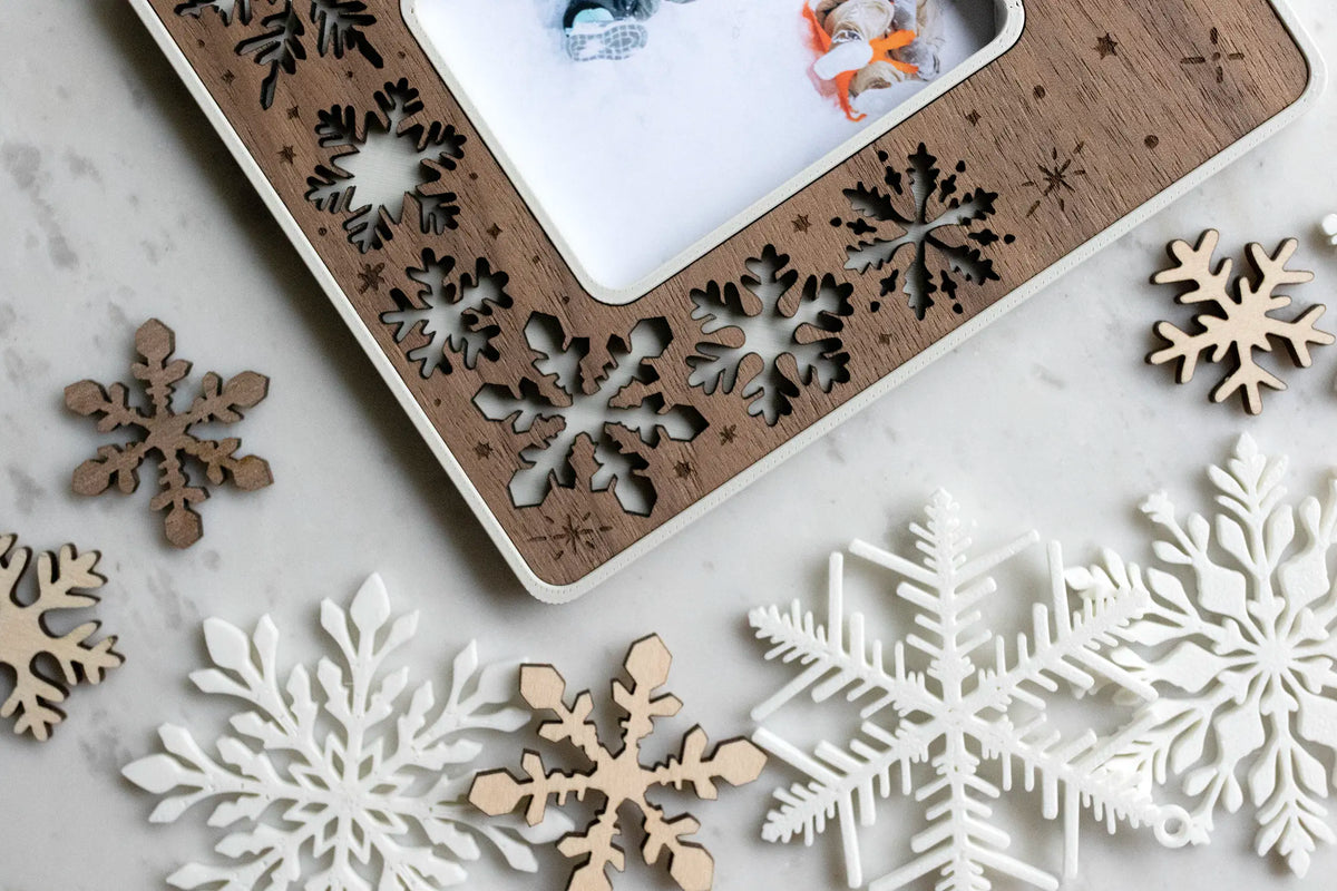 Close-up corner of a laser-cut walnut snowflake magnetic picture frame on white marble, surrounded by scattered wood and white snowflake cutouts, framing a small photo. This holiday keepsake frame is magnetic to stick on the fridge or any magnetic surface.
