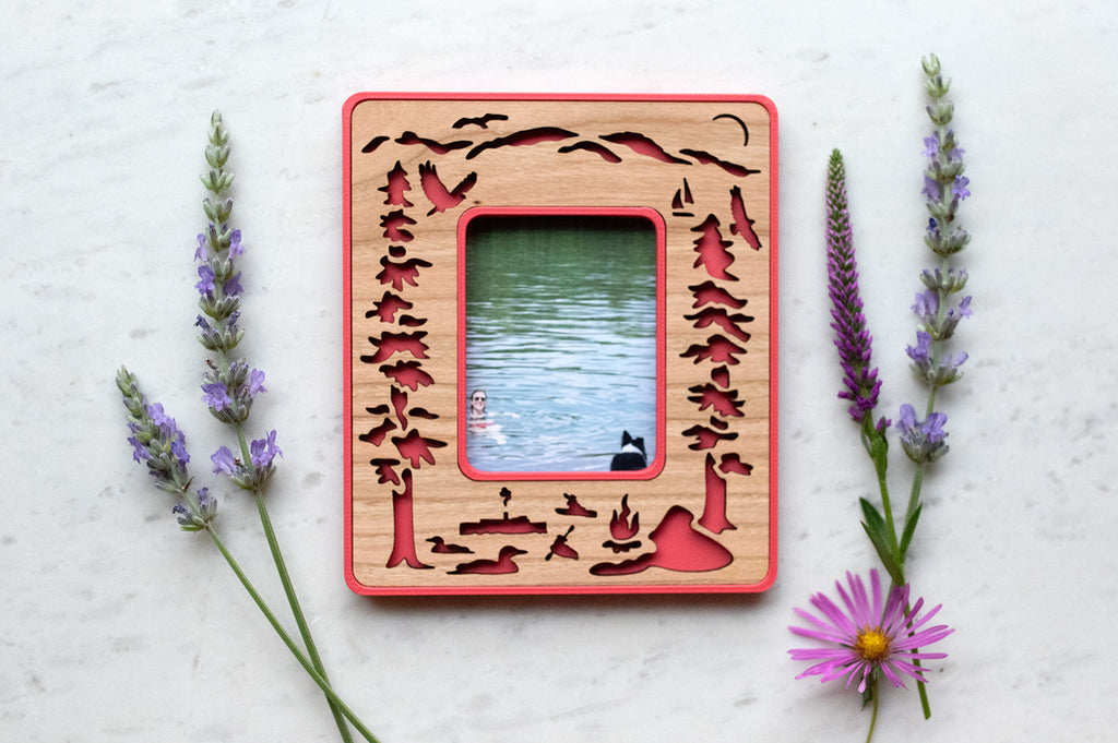 Instax Mini Polaroid photo frame in pink with a laser cut cherry wood inlay, featuring Lake George loons, osprey, and mountains. The frame is surrounded by wildflowers and lying on a white surface. Mini frame displays a photograph of a woman and a dog swimming in Lake George.