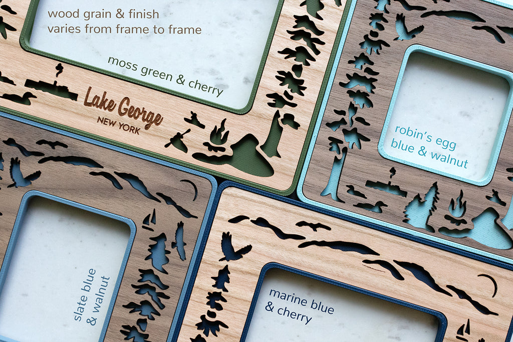 A close-up, detail view of four mini frames with Lake George designs in various shades of blue and green and various wood types. The top left corner of the image reads "wood grain and finish varies from frame to frame". The frames are designed for lake house decor.