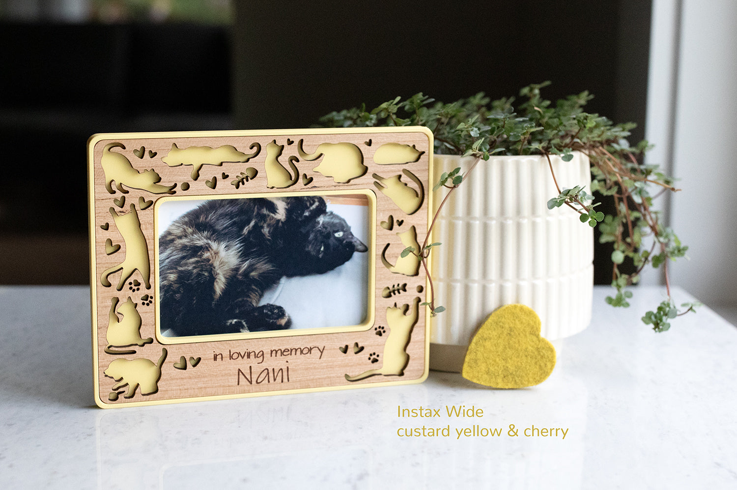 Instax wide magnetic cat memory frame for cat loss gift. The frame is yellow with a cherry wood laser cut inlay. It is propped up next to a white planter and a small yellow felt heart.