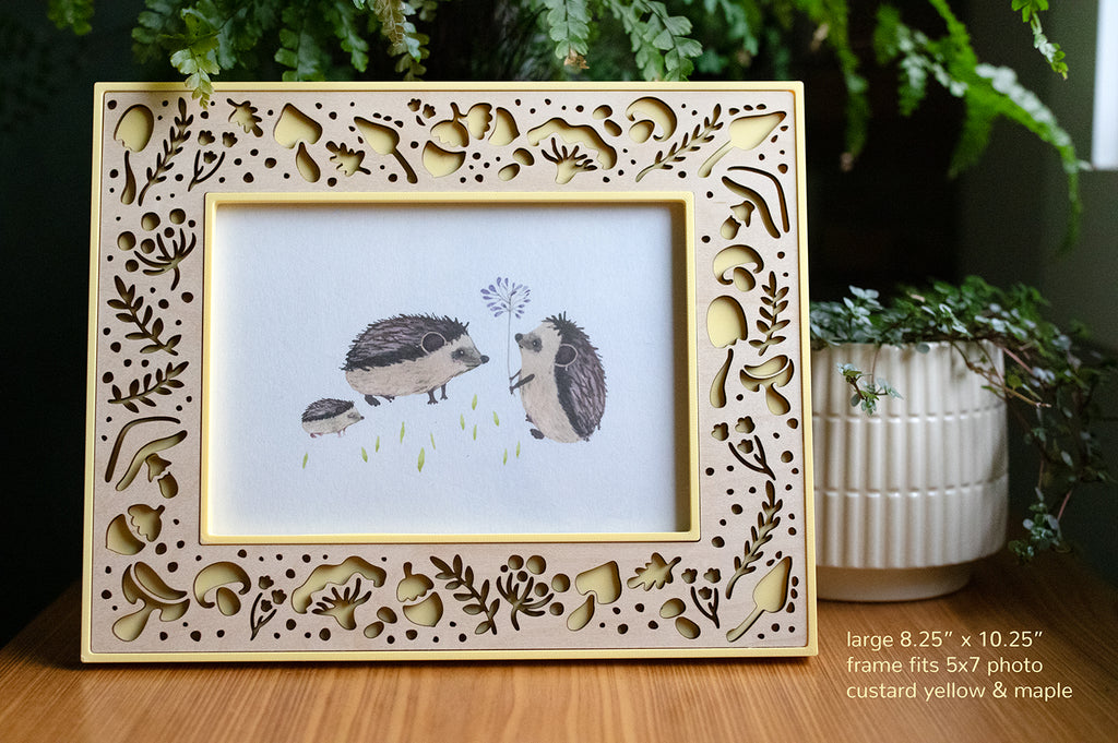 A large 8.25" x 10.25" picture frame that fits a 5x7 photo in horizontal orientation. The frame is pale yellow with a laser cut maple wood inlay. The frame features whimsical mushrooms and acorns. The 5x7 frame displays a photo of a family of hedgehogs, and there is greenery in the background.